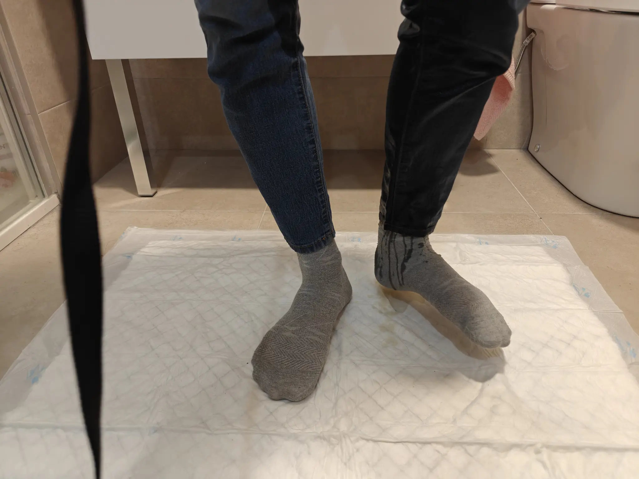 A close-up picture again, this time more focused on my feet. I'm lifting my left foot a little bit, showing its side. The left leg of my pants is completely soaked, and there's trails of pee visible in my gray sock. The right leg is still mostly dry, with a stain starting to show up at the top.