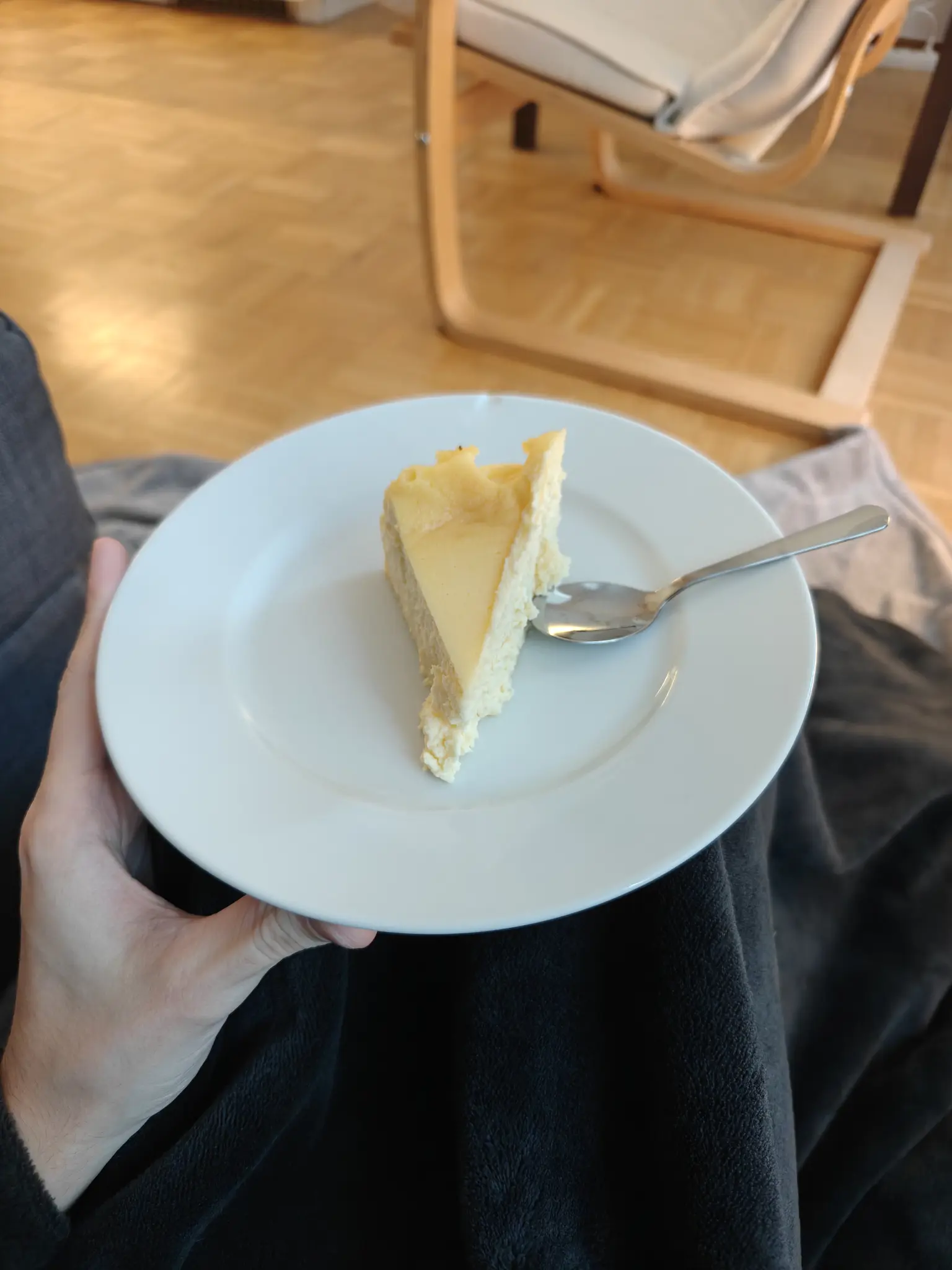 pov picture of me sitting in the floor, covered by a blanket, and holding in my knees a white dish with a portion of cheesecake and a spoon on it