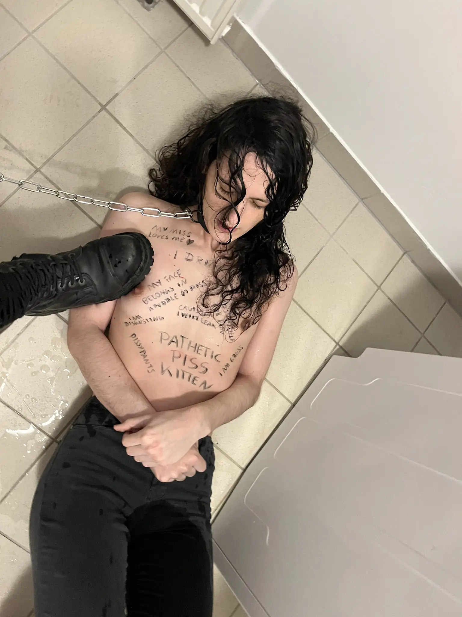 me, shirtless in a bathroom floor, wet from having previously pissed myself, with a chain attached to my collar and being held by my Miss, while she is stepping on my right arm and a bit of my chest with her black boots. I have a lot of different things written on my body with a marker, like "pathetic piss kitten", "pissypants", "I am disgusting", "my face belongs in a puddle of piss", "caution: I will leak", "I piss myself for fun", "I am gross", "I drink piss", and "my Miss loves me 🖤"