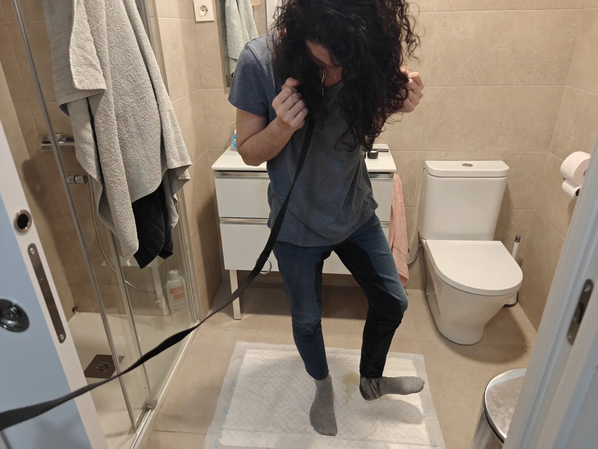 A similar far-off picture. This time I'm no longer covering my face, and I'm lifting my left foot showing it's side. I'm looking down at my legs and the floor, observing how wet they are and how much of a mess I made. The lower half of my left leg is covered in piss, and there's also some trails in my sock. There's a small yellow puddle visible where my left foot was.