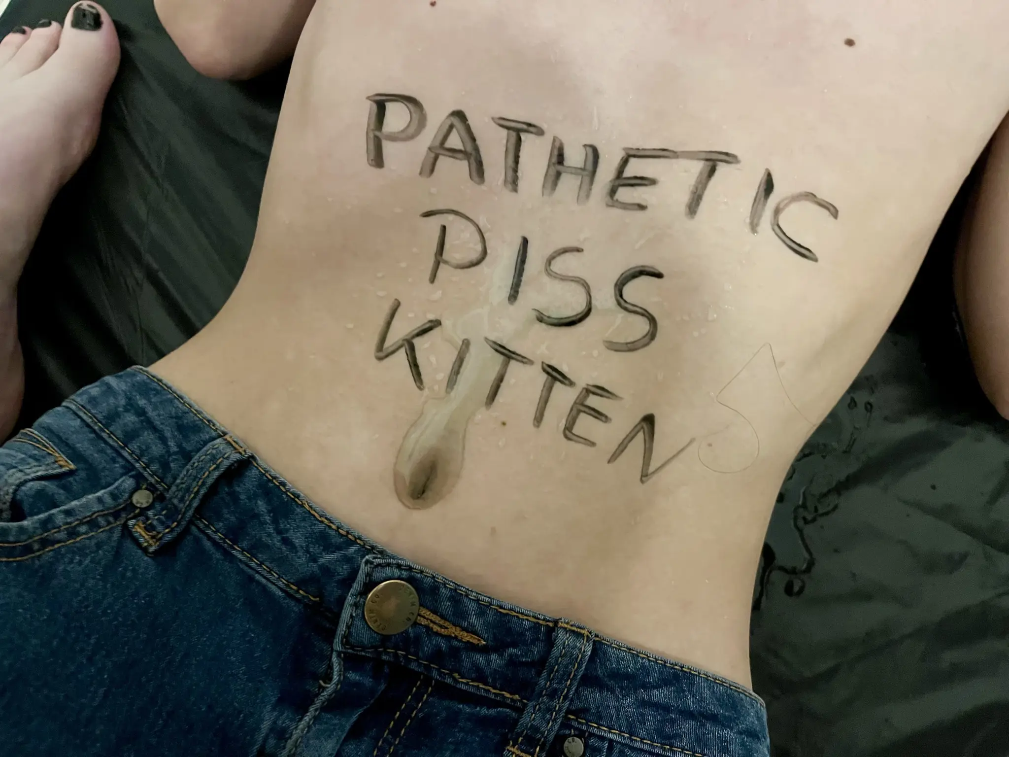 close-up of my wet tummy, where I have "pathetic piss kitten" written with a black body marker, and with piss puddling on my belly button