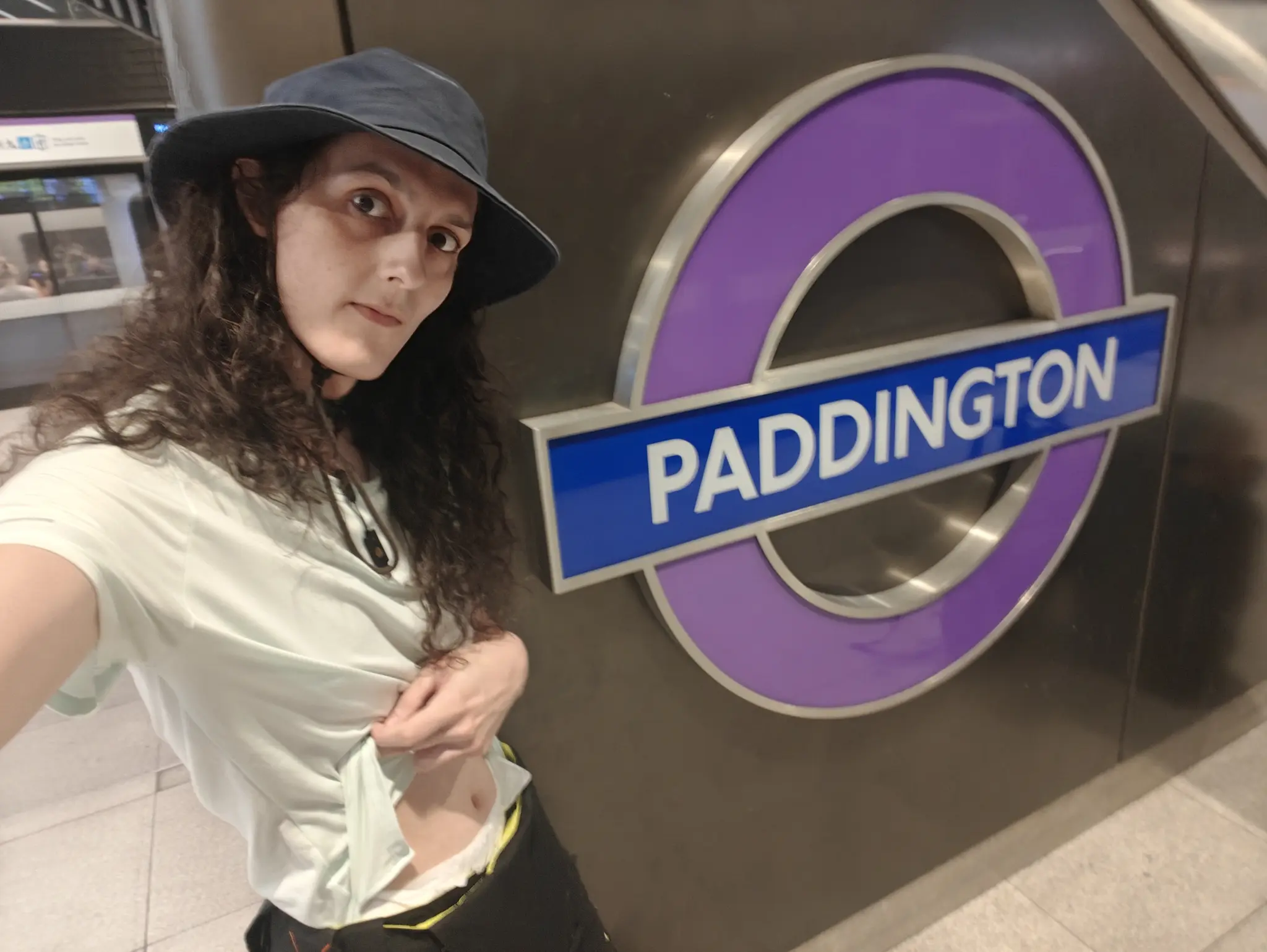 kitten, wearing a blue hat, bright lime t-shirt, and overalls to their waist, in front of the Elizabeth's line sign for Paddington station. they're lifting their t-shirt slightly, revealing a peeking white diaper