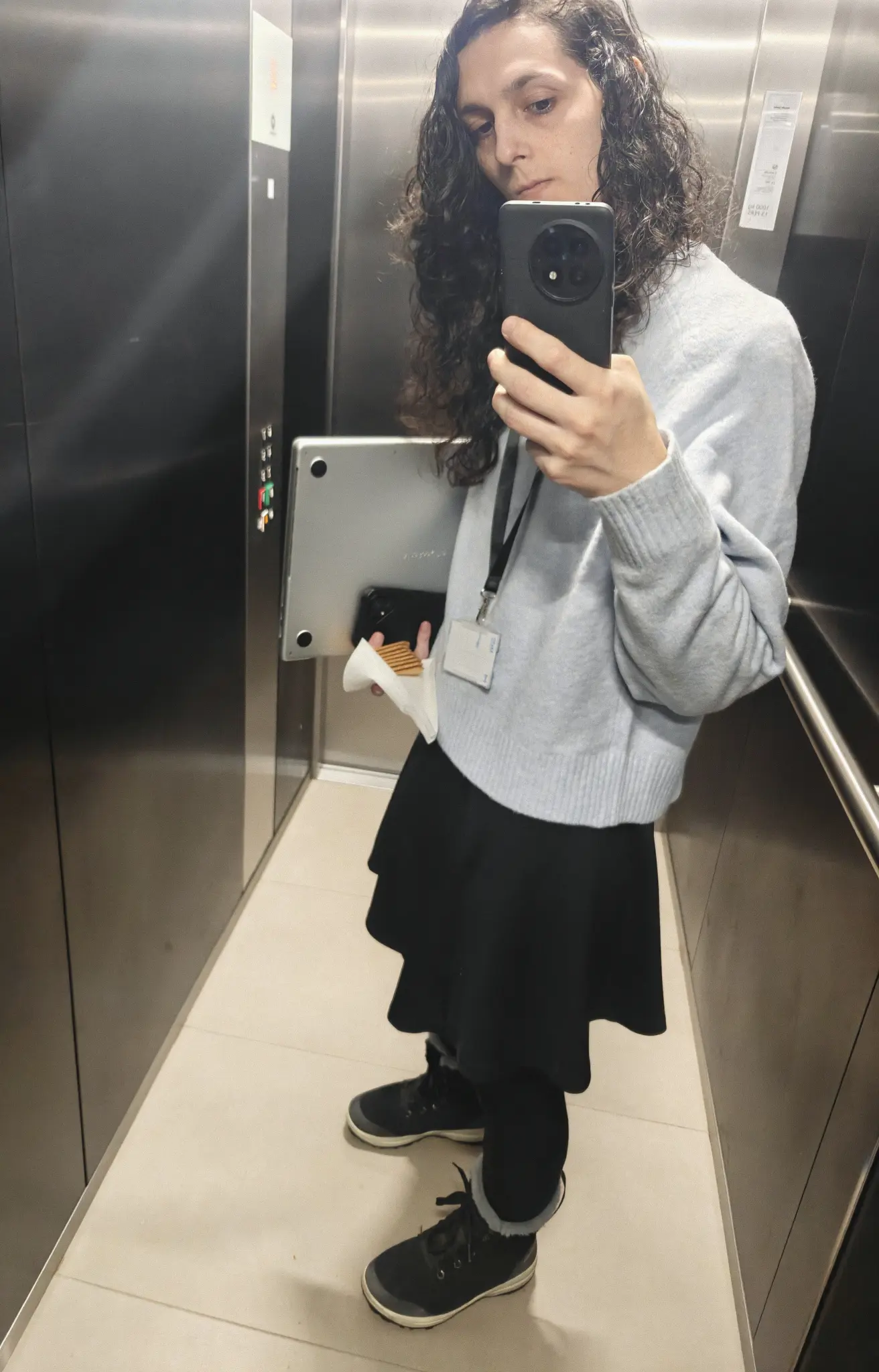 kitten taking a selfie in an elevator, wearing a light blue sweater, a black skirt, and black stockings, while carrying a laptop, a second phone, and a stack of biscuits