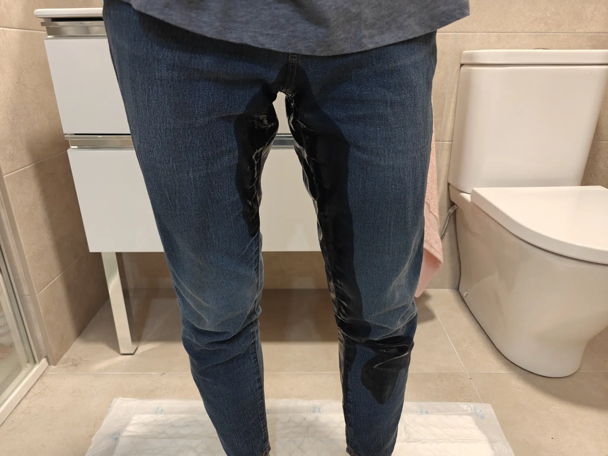 Close-up picture of my legs, while I'm standing in my bathroom, on top of a slightly visible waterproof mat, and with the toilet and the sink in the background.

There's a glistering stain of pee in my jeans coming from my crotch, going down both legs, but specially the left leg, where it has already gone past my knee.