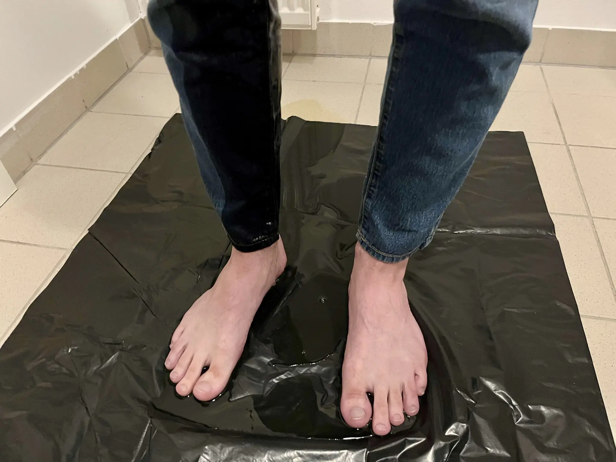 Close-up of the lower part of my legs, showing the right leg of my jeans soaked, and my feet on top of a yellowy puddle that has went beyond the black plastic bag we put on the floor